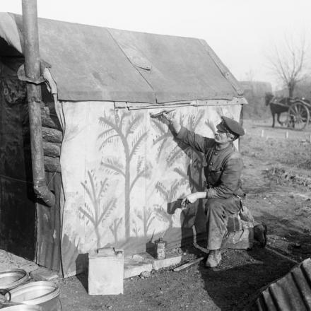 Un soldato del Corpo dei servizi dell'esercito (ASC) dipinge alberi su un telo impermeabile che copre il lato della sua capanna per mimetizzarla, vicino a Boesinghe, il 28 gennaio 1918. © IWM Q 10634