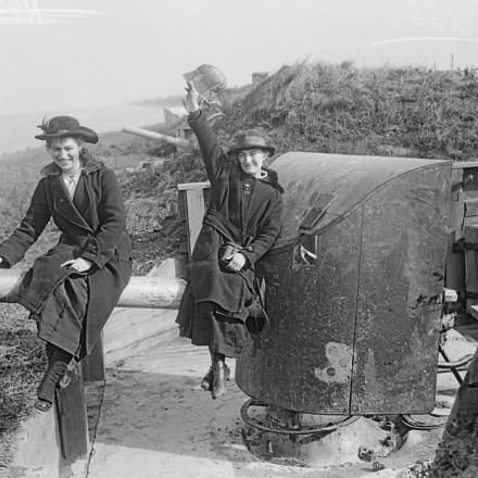 Due ragazze belghe sedute sulla canna di un cannone tedesco di difesa costiera alla Eylau Battery, Ostenda, 19 ottobre 1918. Una ha un elmetto tedesco dipinto di camouflage (Stahlhelm) e indossa una spilla di bandiere alleate. © IWM (Q 11380)
